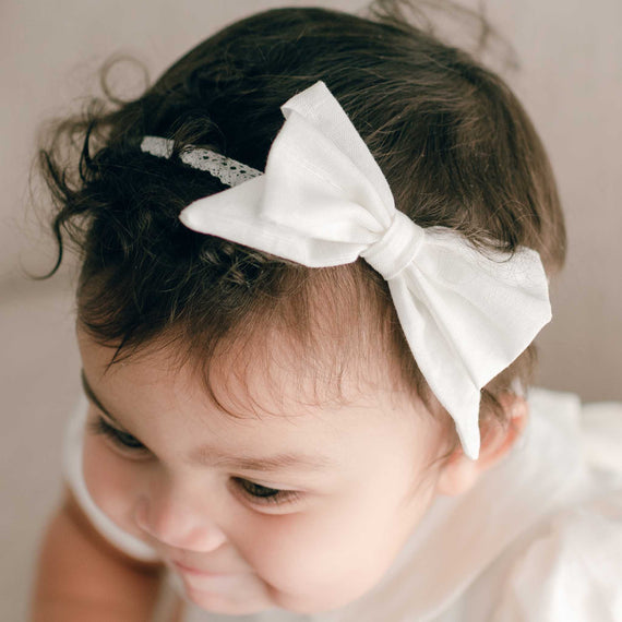 A close-up photo of a baby with curly hair wearing an Emma Linen Bow Headband, looking downward with a gentle, thoughtful expression.