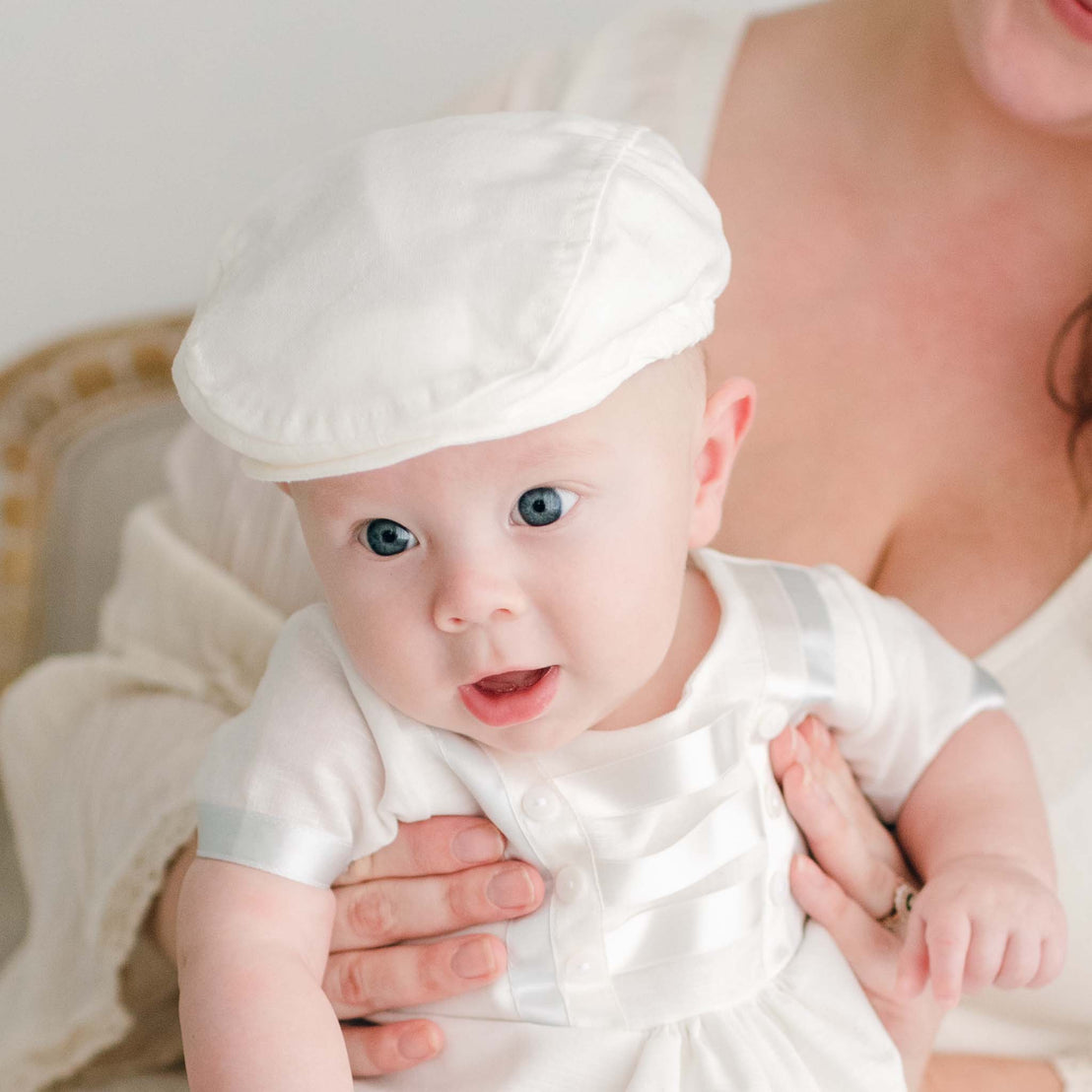 A baby with bright blue eyes wearing an Owen Romper Accessory Bundle sits cradled in the arms of an adult, glancing slightly to the side.