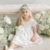 A young girl sitting on the floor wearing the Lola Girls Lace Dress with a white floral crown and holding a bouquet of white flowers, surrounded by white roses.