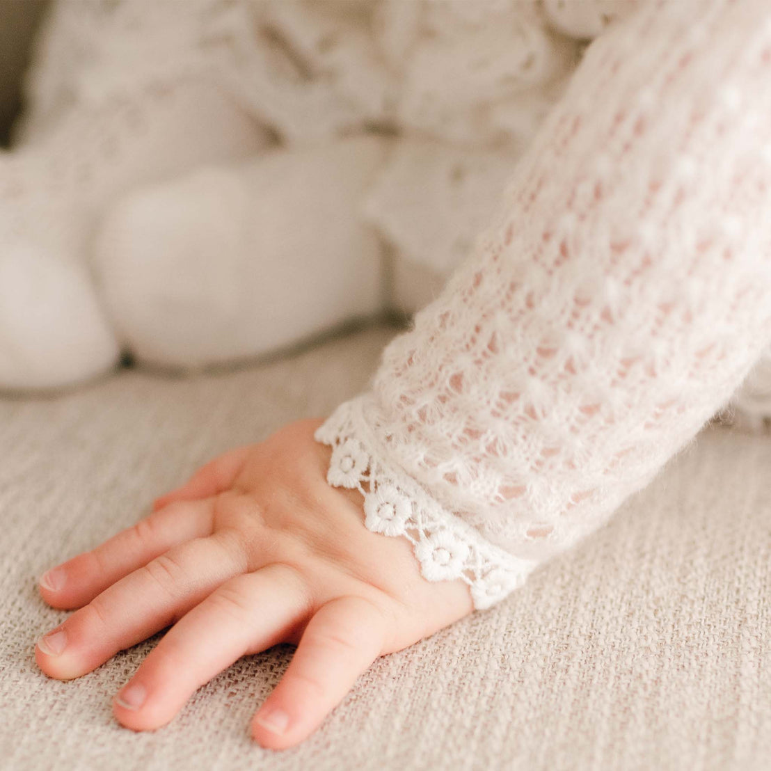 Detail of the Lola Knit Sweater sleeve. Baby girl wearing the Lola Knit Sweater that matches the Lola Christening Gown, Dress and Romper. Made of 100% ivory knit acrylic in light ivory, a soft and breathable knit with button closures and light ivory trim.