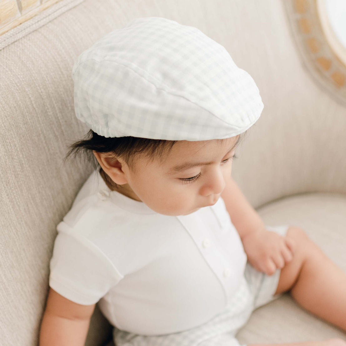 A baby wearing an Ian Checked Newsboy Cap and white outfit, sitting on a beige cushioned chair, looking down thoughtfully.