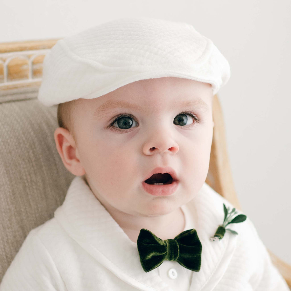 A baby with bright blue eyes and an open mouth wears a white christening outfit with a green bow tie and the Noah Ivory Newsboy Cap, sitting against a light background.