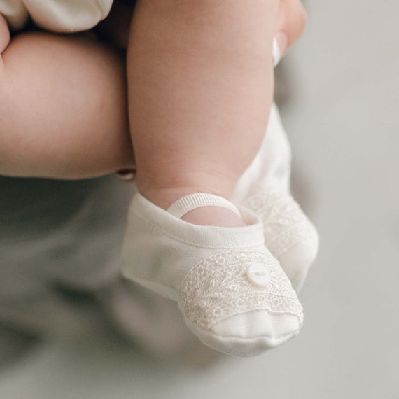 The top detail of the Oliver Booties in ivory worn by a baby boy. The Oliver booties are made with a linen top, soft pima cotton bottom, and features a Venice lace and button detail (with elastic strap across the foot).