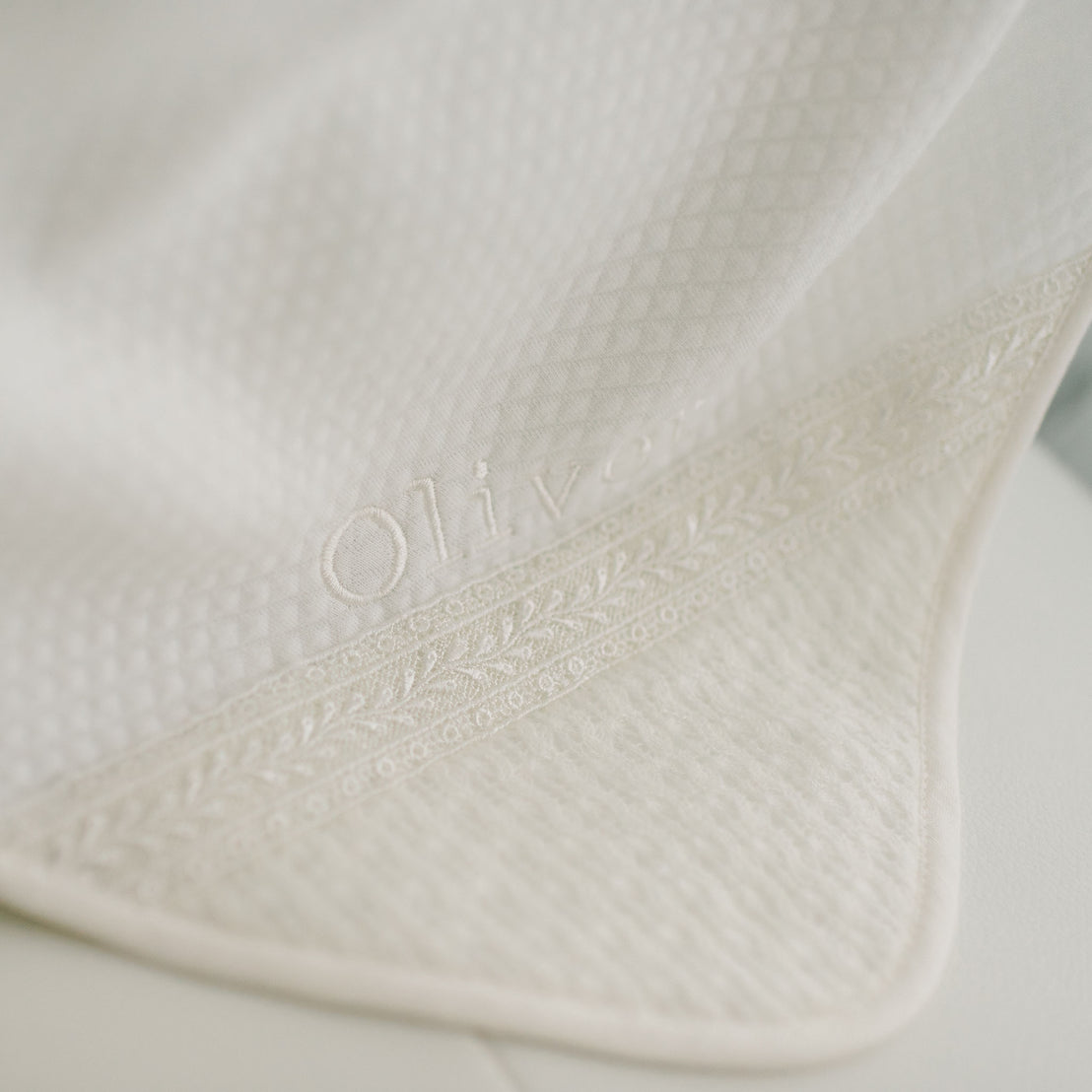 A close-up image of the ivory Oliver Personalized Blanket featuring the ivory, textured cotton fabric with delicate embroidered detailing. The embroidery includes the name "Oliver" and intricate, leafy patterns along the trim.