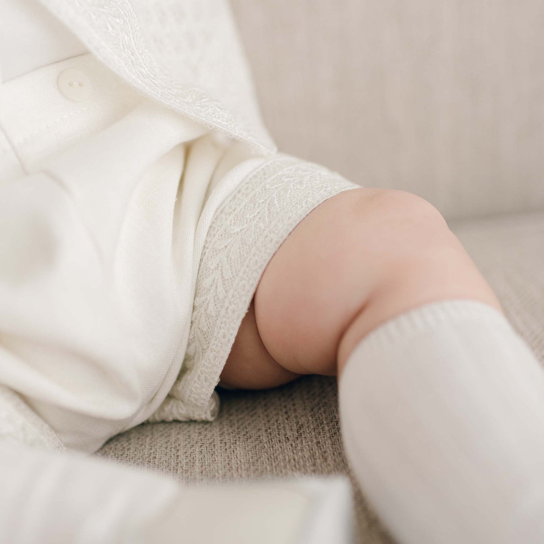 Close-up image of a baby's leg and foot, showing them wearing the light ivory Oliver Shorts. The baby is wearing the light ivory Oliver Sweater Shorts Suit Set with Venice lace lace along the hem of the shorts and sweater.