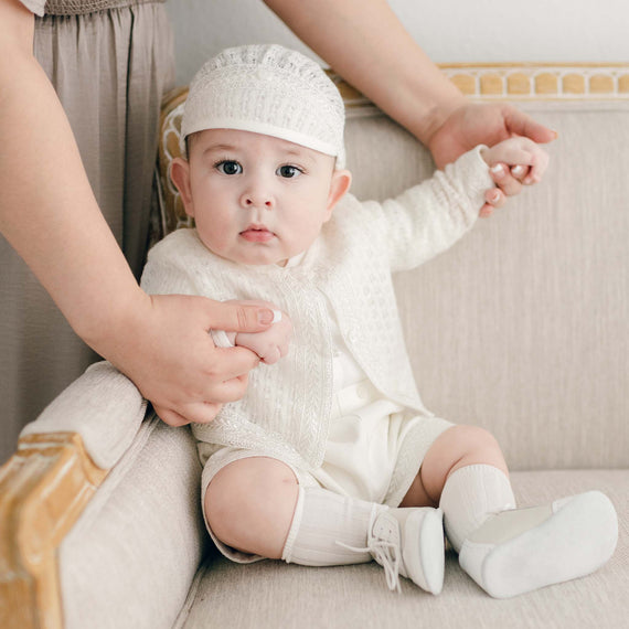 A baby boy in the light ivory Oliver Sweater Shorts Suit sits on a beige sofa, held securely by a parent's hand. The baby boy, wearing an ivory Oliver Knit Hat and Boys Ivory Two Tone Wingtip Shoes, looks curiously at the camera.