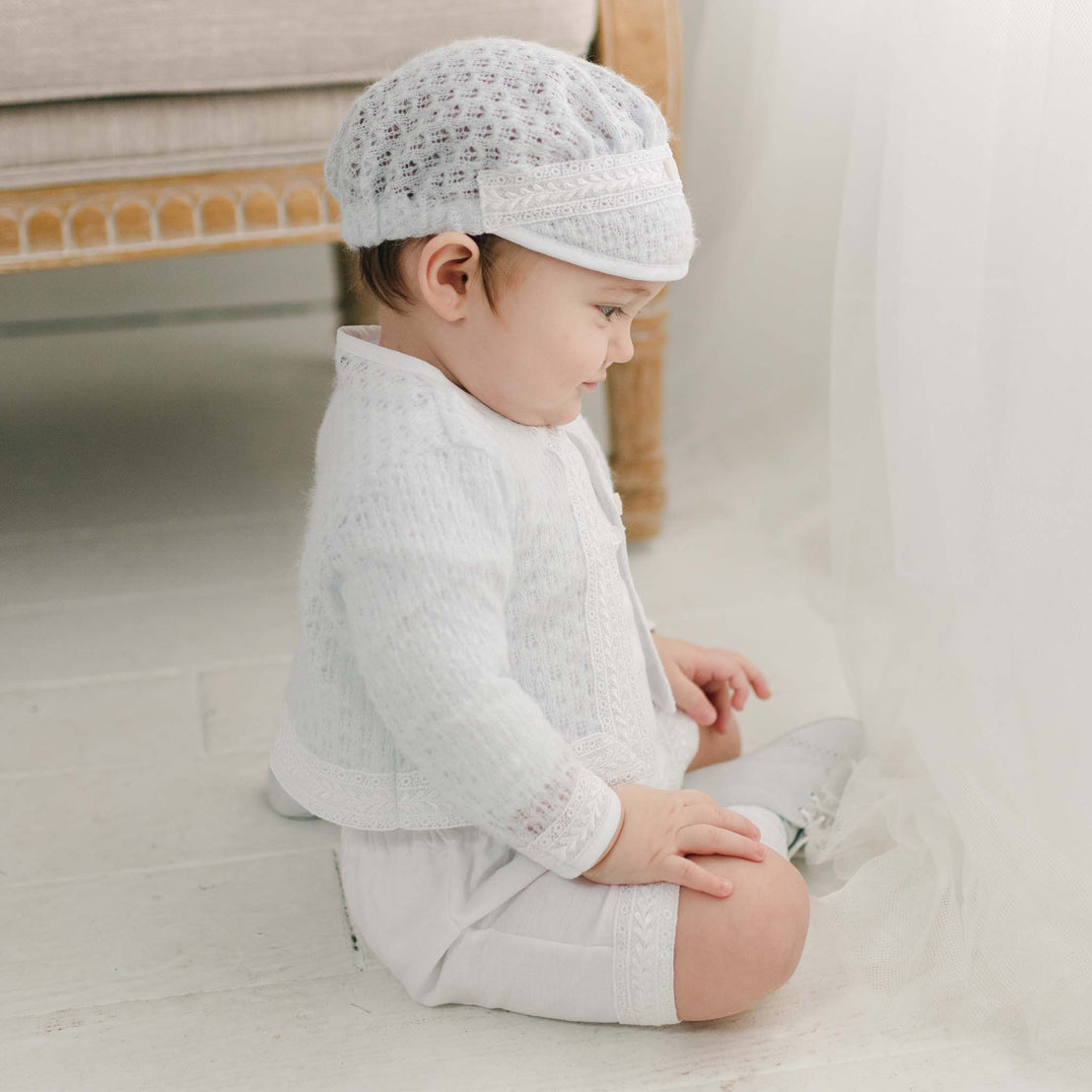 A toddler sitting on the floor, wearing the light blue knit Oliver Sweater Shorts Suit and cap, gently touching a sheer curtain with a soft, focused expression.