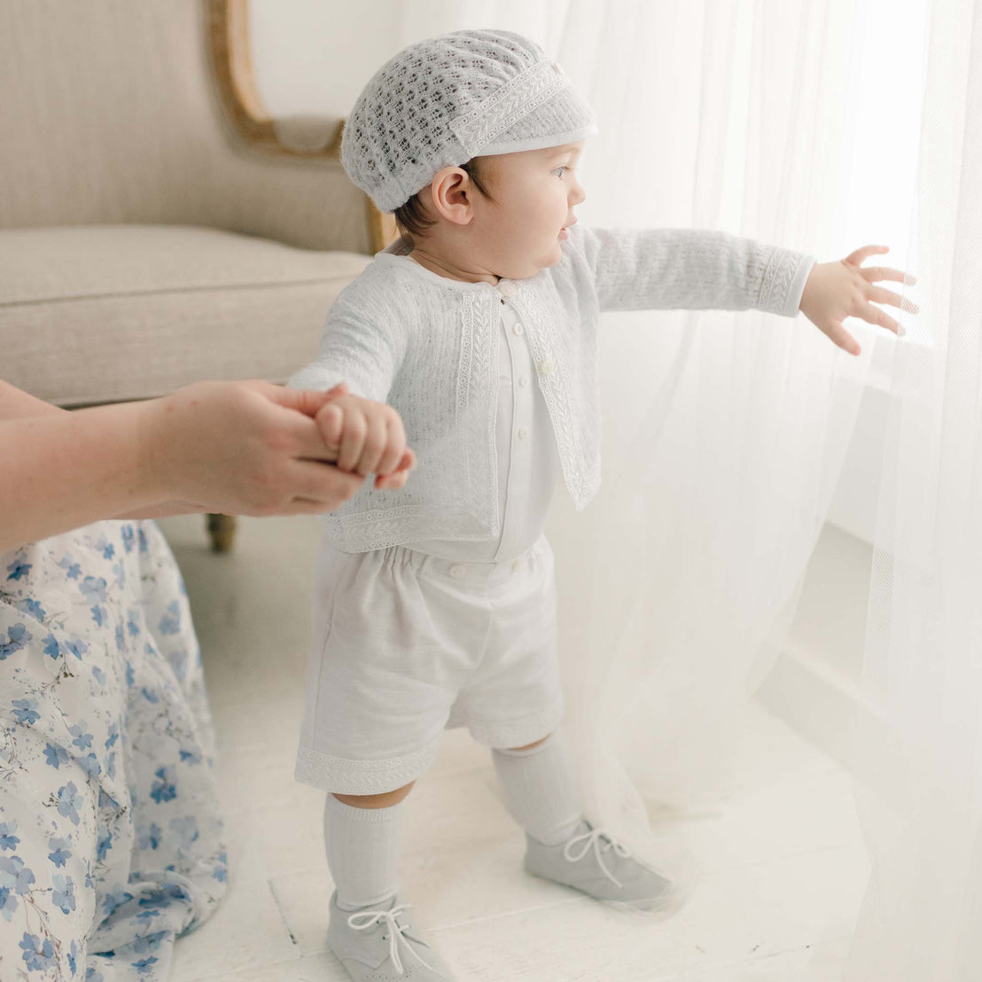 A toddler in the light blue Oliver Sweater Shorts Suit takes tentative steps while holding an adult's hand, beside a sheer curtained window in a softly lit room.