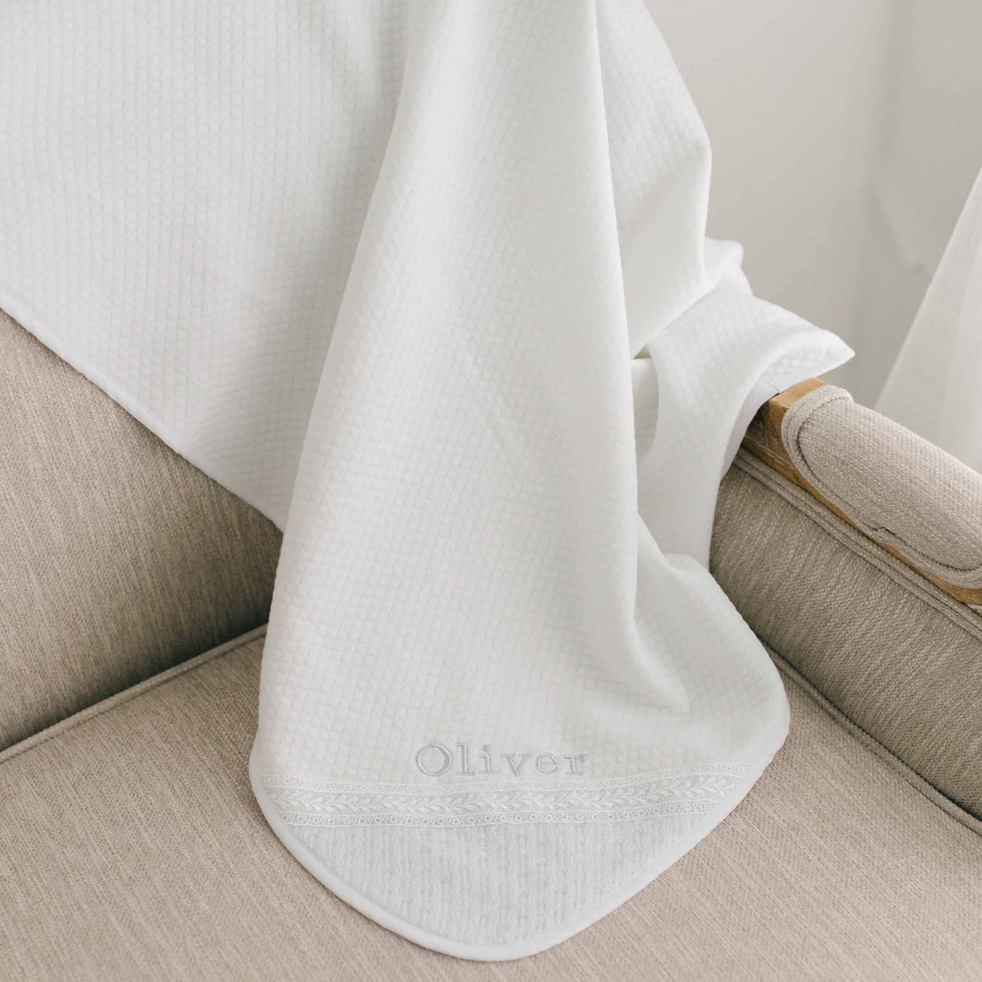 A white Oliver Personalized Blanket drapes over the arm and seat of a light beige chair. The embroidery includes the name "Oliver" and intricate, leafy patterns along the corner of the blanket.