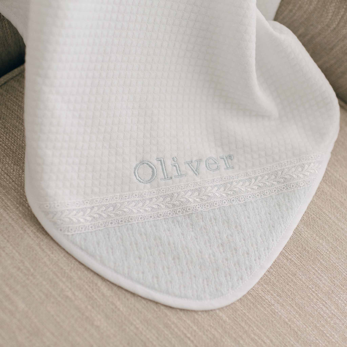 A close up of the white Oliver Personalized Blanket draped over a beige couch. The bottom edge of this personalized heirloom features delicate lace detailing and is embroidered with the name "Oliver" in light blue thread.