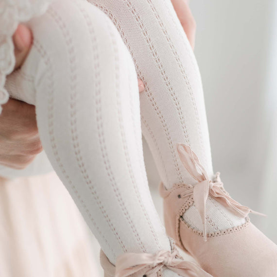 Close-up of a person adjusting a pink ballet slipper on their foot, highlighting the detailed stitching and Openwork Fancy Tights against a pale background.