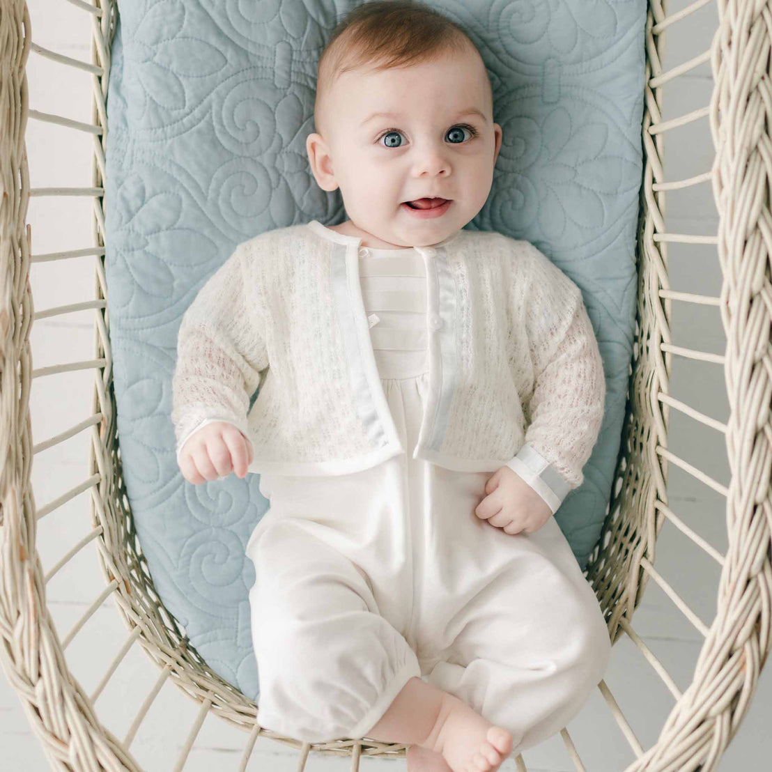 A joyful baby with blue eyes and light hair, wearing an Owen Knit Sweater adorned with a blue silk ribbon, lies in a woven basket, looking up with a bright smile.