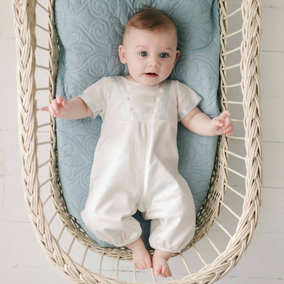 A baby with blue eyes lies comfortably in a wicker basket, dressed in the Owen Linen Romper, against a soft blue patterned cushion background.