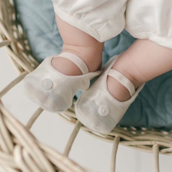 Close-up of a baby's feet in the Owen Linen Booties, resting on the edge of a wicker basket lined with a soft, blue cushion.