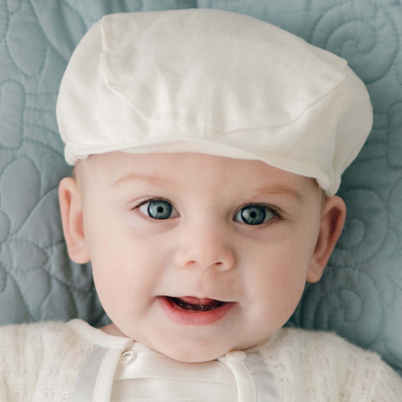 A baby with bright blue eyes and a joyful smile, wearing the Owen Linen Newsboy Cap, on a light blue textured background.