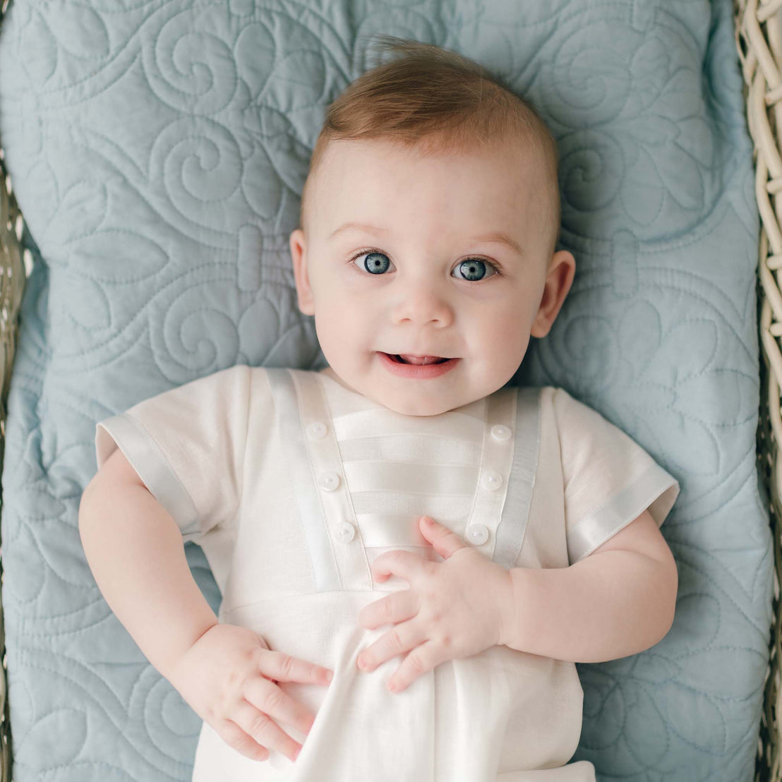 A smiling baby with bright blue eyes lies on a quilted blue blanket, dressed in the Owen Linen Romper, looking directly at the camera. The romper features silk ribbon in ivory and light blue across the front bodice and sleeves.