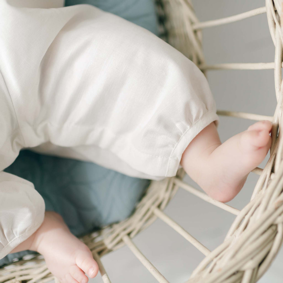 A close-up image of a baby's feet and legs clad in a soft white Owen Linen Romper, folded and poking through a woven basket on a light background.