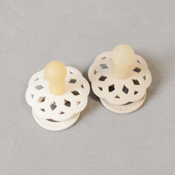 Two Grace Pacifiers | Ivory & Vanilla colored pacifiers with round shields and beige nipples, manufactured in Denmark, placed on a light grey textured surface.