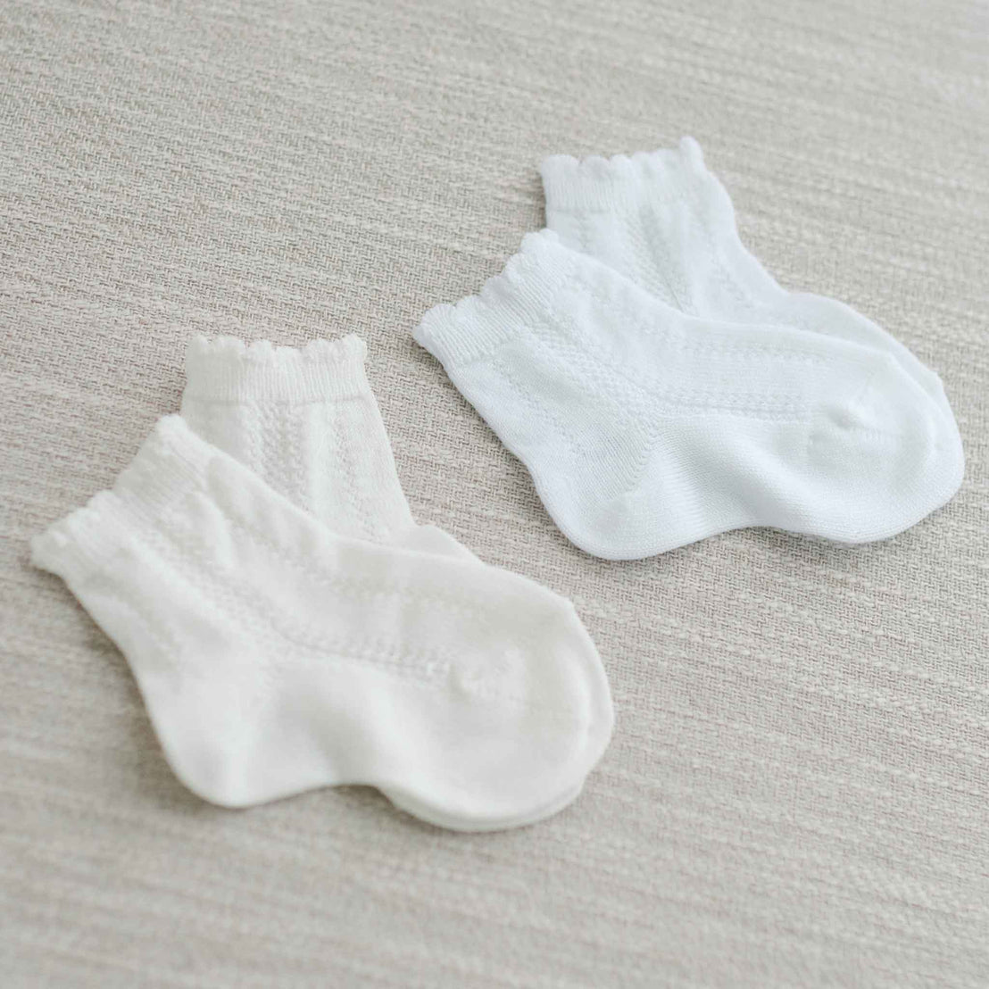Two pairs of small white Pattern Socks with scalloped cuffs are laid flat against a light beige textured fabric surface.