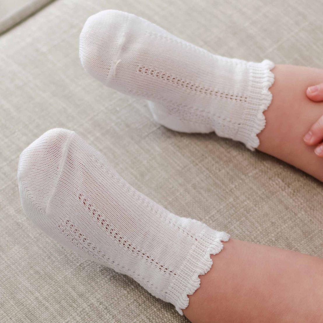 A close-up image of a child's feet wearing white, cotton Pattern Socks against a soft beige background. The socks feature a delicate lace pattern along the scalloped cuff.