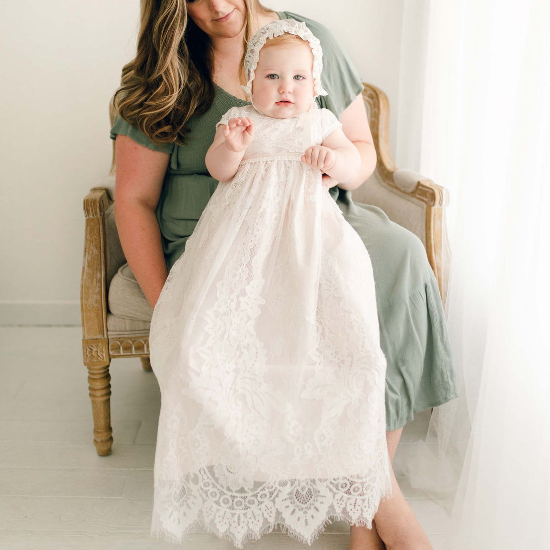 Mother with a baby girl wearing the pink Victoria Puff Sleeve Christening Gown and Bonnet. The gown is made with ivory embroidered lace over a pink silk Dupioni lining.