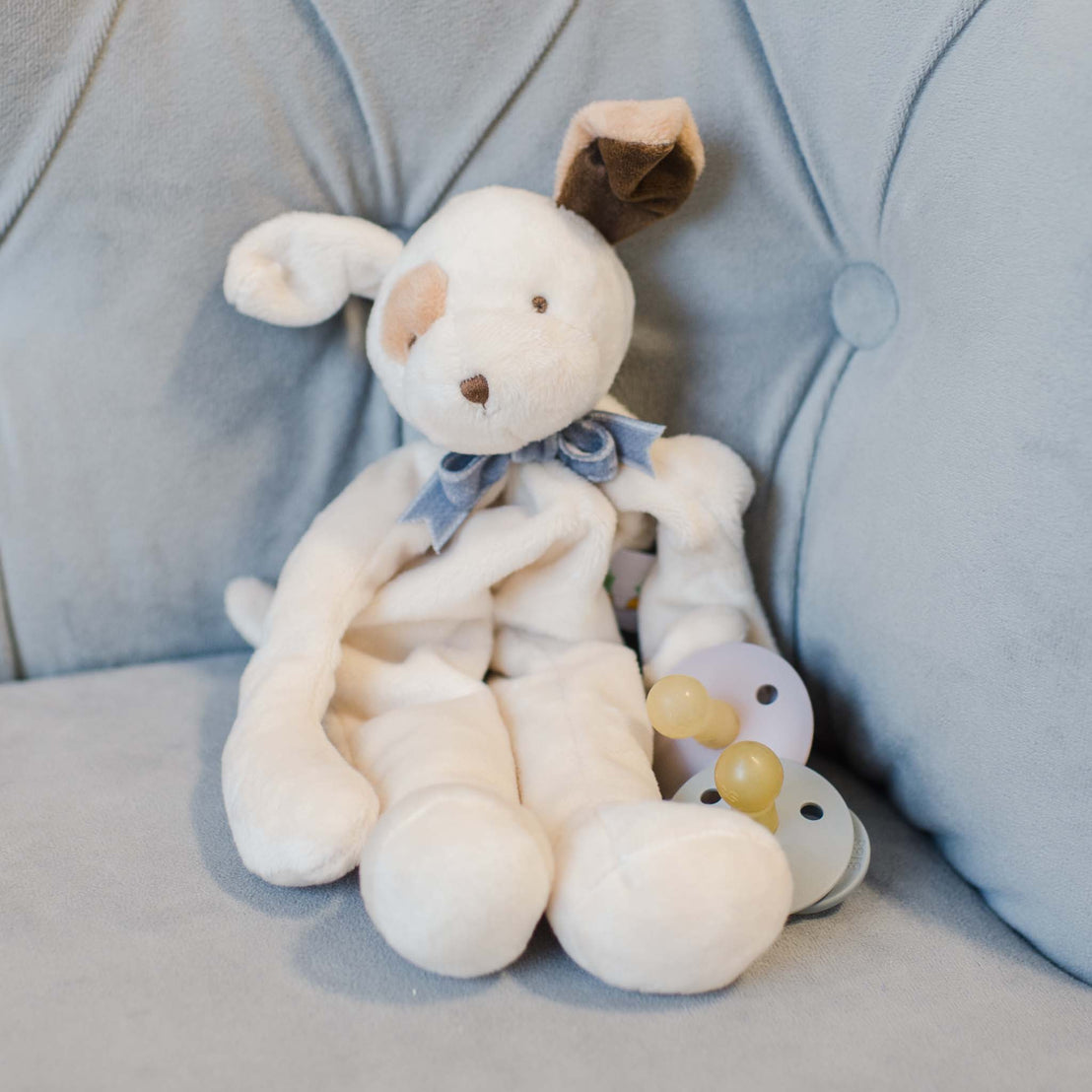 The Ezra Heather Silly Puppy Buddy stuffed animal sitting on a couch. This stuffed animal serves as a pacifier holder