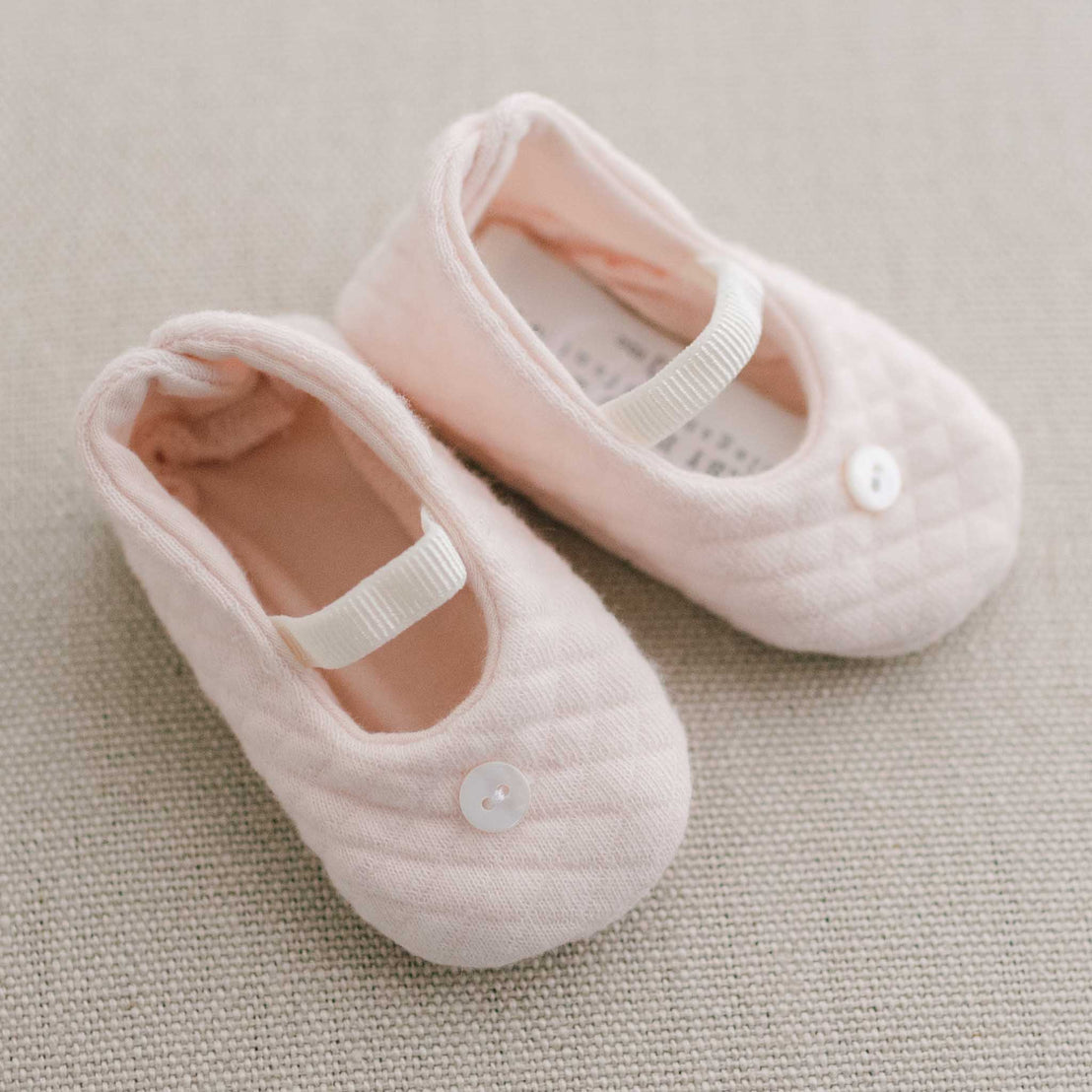 A pair of Ava Quilted Booties in blush pink, each adorned with a small white button, resting on a soft grey textured background.