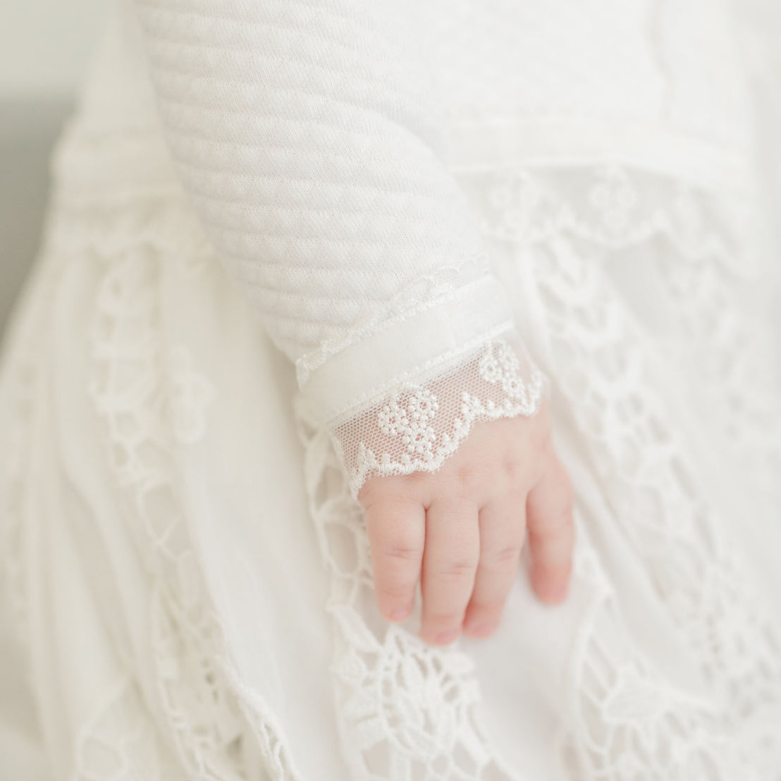 Close-up of a baby's hand against a Grace Quilted Cotton Sweater with floral lace details, featuring a soft sleeve edge and pearl style buttons.