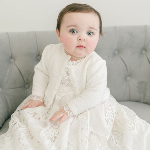A baby with big blue eyes, wearing a white floral lace dress and Grace Quilted Cotton Sweater with pearl style buttons, sitting on a gray tufted sofa.
