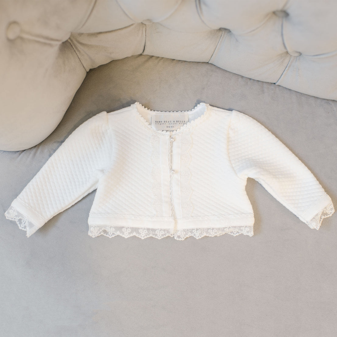A small white Grace Quilted Cotton Sweater with floral lace trim is laid out on a soft gray surface.