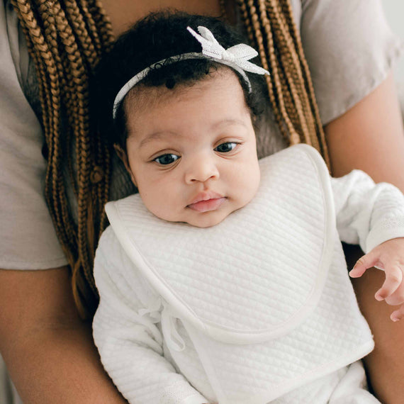Baby girl with her mother. The baby wearing the color coordinated Ivory Quinn Bib. The bib is made with classic quilted textured cotton with matching ties and trim in soft pima cotton.