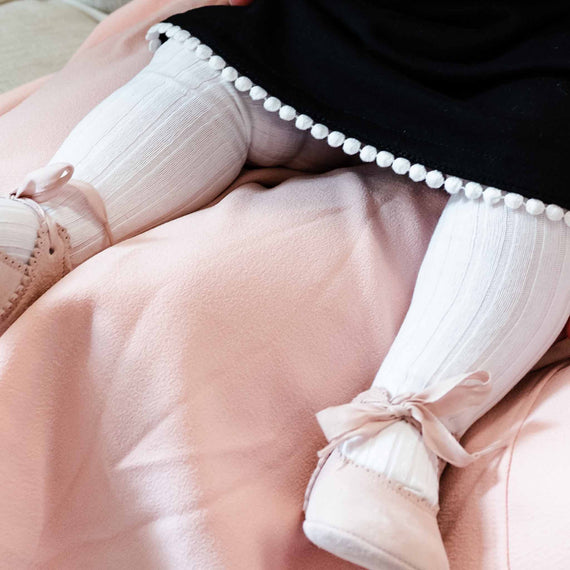 A close-up view of a baby sitting on a pink blanket, wearing white pleated pants with a black top decorated with white pompoms, and pink shoes with bow details made in Spain.