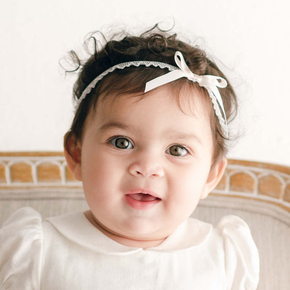 A cheerful baby with curly hair wearing a boutique white dress and the Emma Pink Silk Ribbon Headband, smiling while sitting in a vintage-inspired chair against a neutral background.