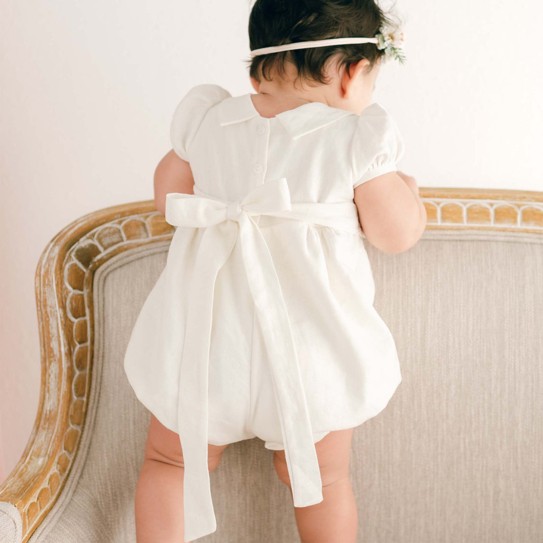 A baby in an Emma Bubble Romper with a bow on the back stands facing away from the camera, leaning on an ornate, vintage chair. A delicate floral headband adorns the baby's head.