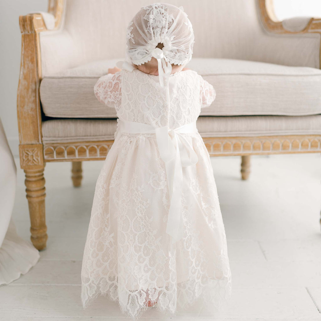 A toddler in a delicate white lace Juliette Romper Dress and a matching headband stands with her back to the camera in front of an elegant sofa in a softly lit, upscale room.