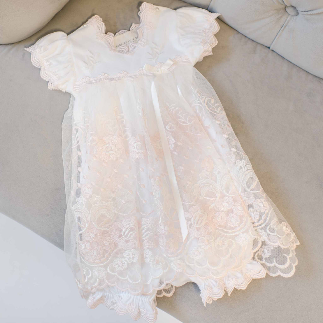 A delicate white Joli Romper Dress with intricate lace detailing and a sheer overlay, displayed on a soft beige sofa, perfect for an upscale boutique.