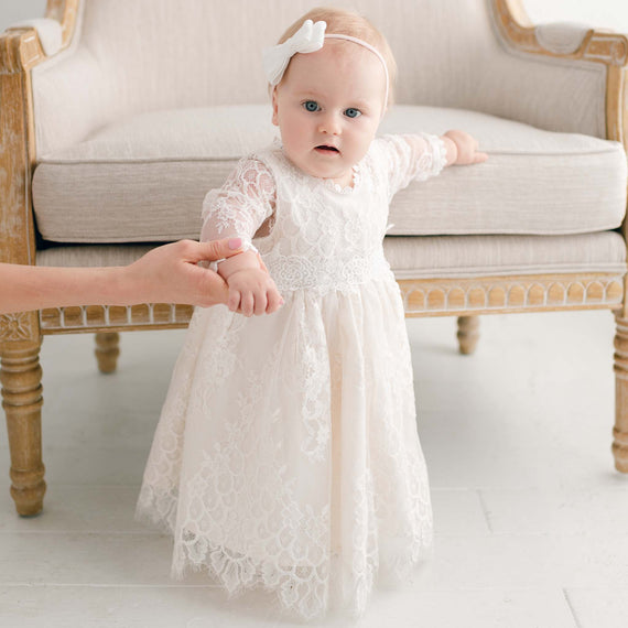 A toddler in a luxury white lace Juliette Romper Dress and headband stands while holding an adult's hand, with a vintage couch in the background.