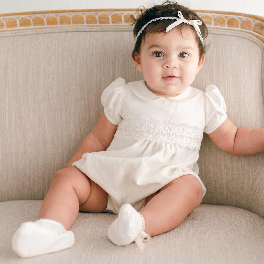 A newborn baby girl with big eyes sits on a beige sofa, wearing an Emma Bubble Romper with lace details and white socks. She has a white headband with a bow.