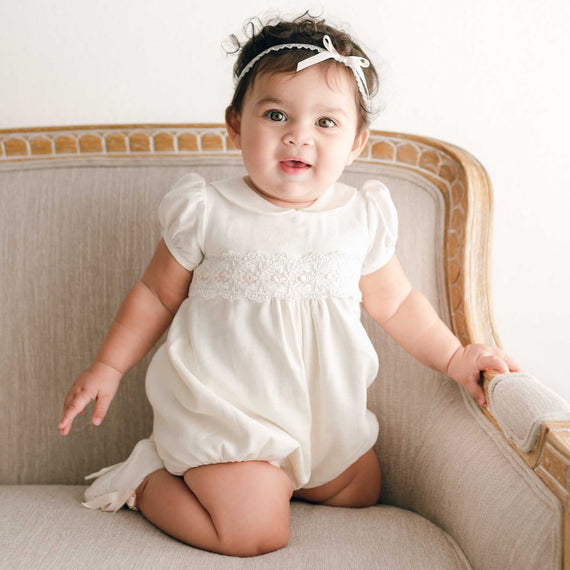 A cheerful newborn in an Emma Bubble Romper smiles while sitting on a beige vintage-inspired sofa, wearing a cute white headband.