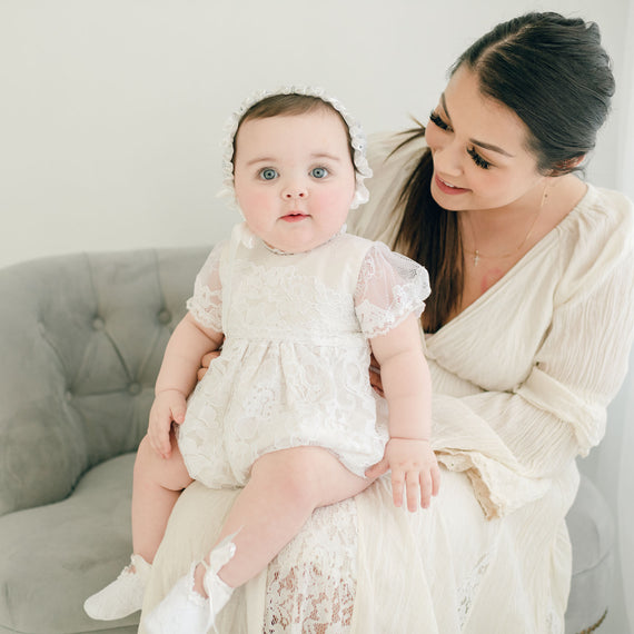 A mother, with dark hair tied back, smiles gently at her baby daughter sitting on her lap. The baby, wearing an Aria Bubble Romper with matching headband, looks towards the camera.