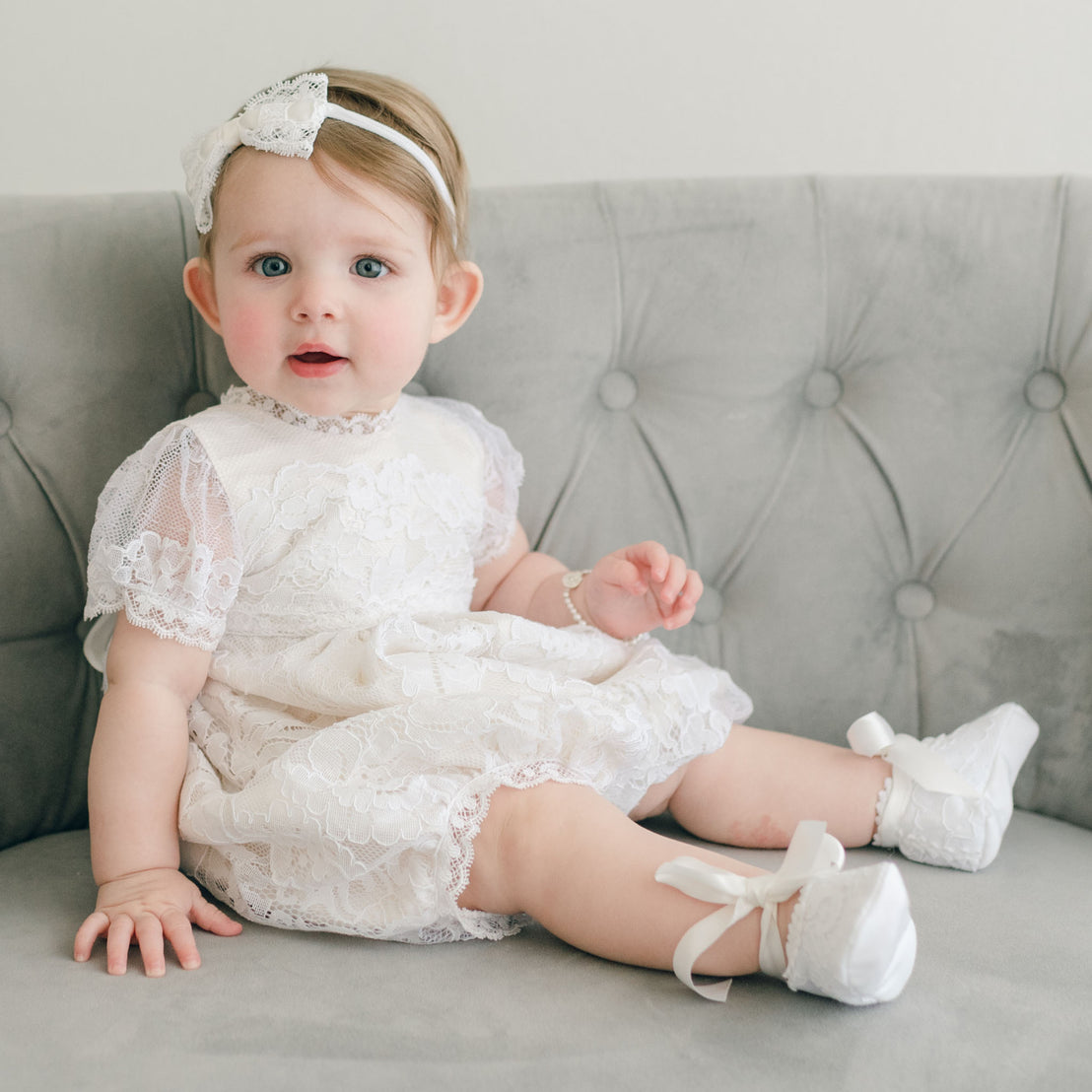 A baby girl with blue eyes, wearing an Aria Bubble Romper and headband, sits on a grey sofa. She is also wearing the Light Ivory Lace Booties.