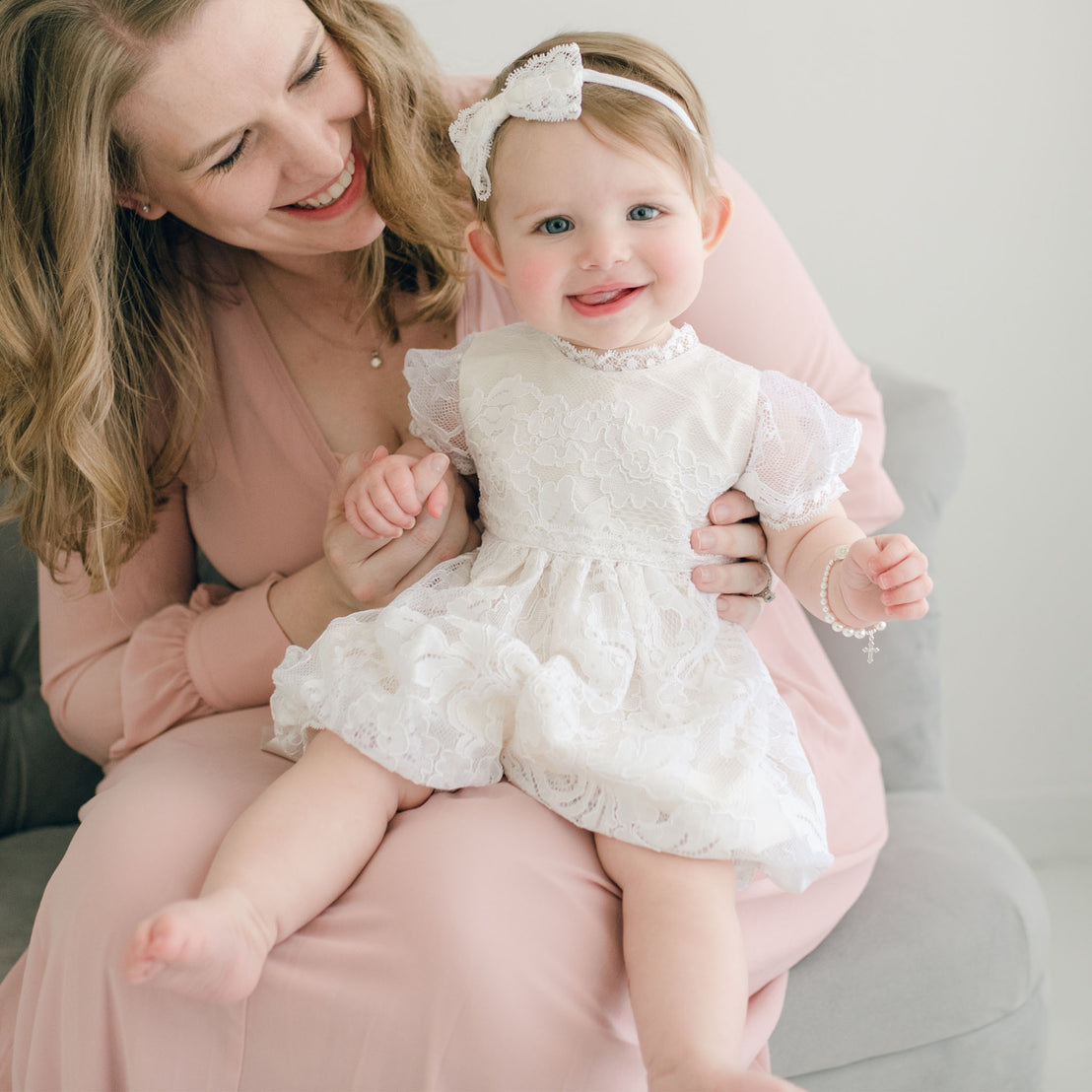 A joyful woman in a pink dress holding a smiling baby girl in an Aria Bubble Romper and Aria Lace Bow Headband, sitting together on a gray armchair in a softly lit room.
