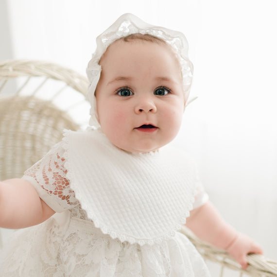 A baby with blue eyes and a Rose Bib of ivory Venice floral lace, with a matching headband, sits in a wicker chair, looking curious and adorable.