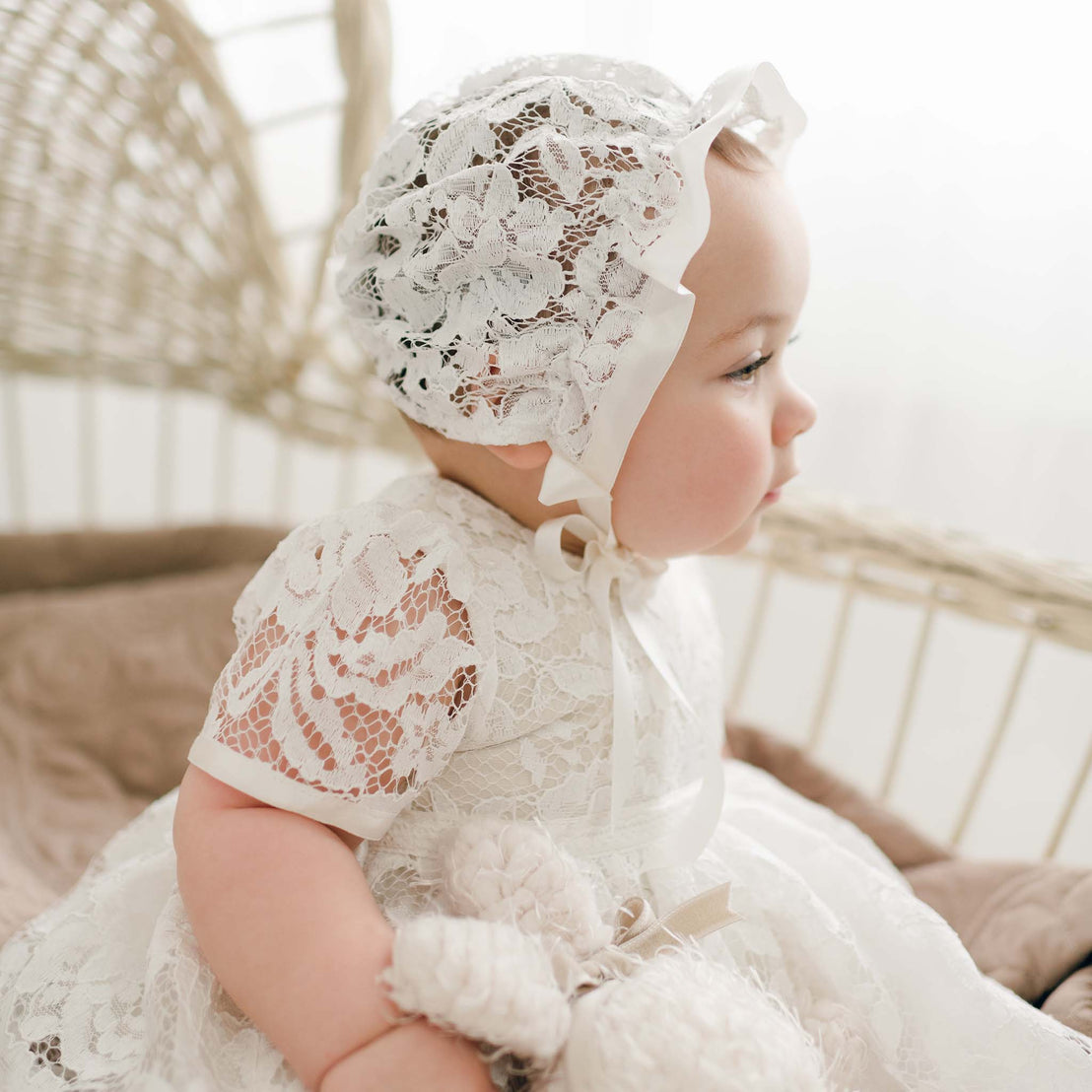 A baby wearing the Rose Lace Bonnet and matching Rose Christening Gown sits in a wicker basket, gazing to the side with a reflective expression. The setting conveys a soft, serene mood.