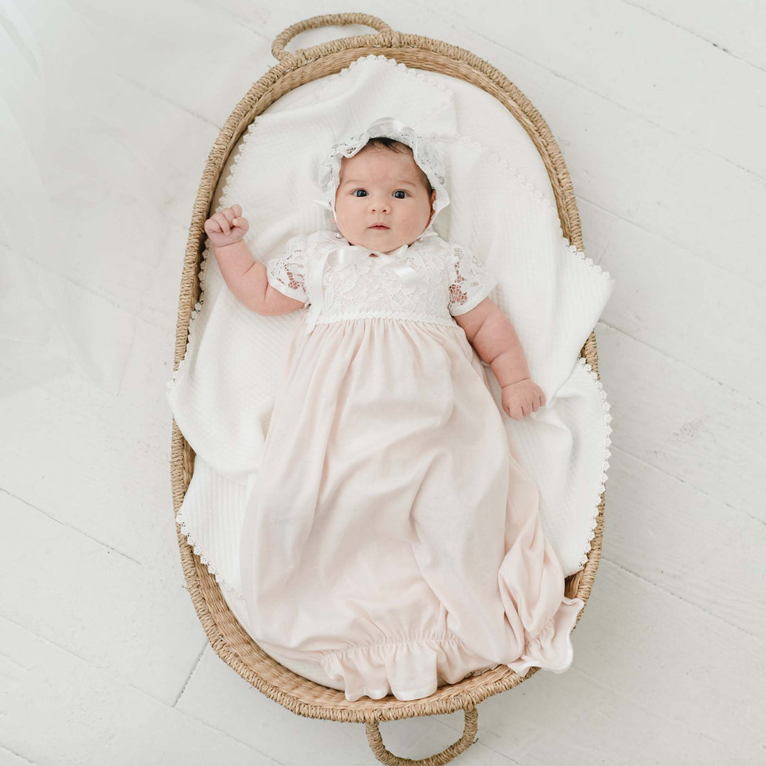 A baby dressed in a Rose Layette & Bonnet and headband lies in a vintage woven basket on a white floor, looking upwards with wide eyes.