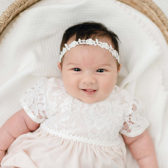 A smiling baby wearing a traditional, heirloom white lace dress and a Rose Lace Headband lies comfortably in a basket covered with an upscale soft blanket.