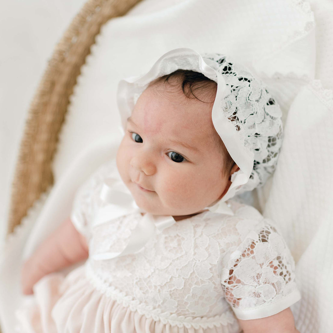 A baby with dark hair and light skin wearing the Rose Layette & Bonnet looks upwards thoughtfully, set against a soft, white background.