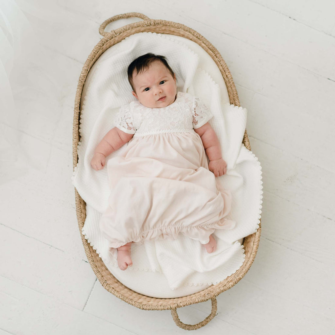 A newborn baby dressed in the Rose Layette & Bonnet lies in an heirloom woven oval bassinet on a white floor, looking directly at the camera.