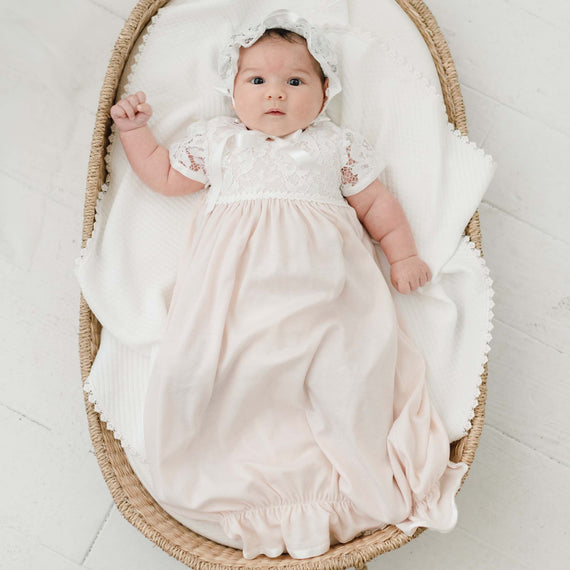 A baby wearing a Rose Layette & Bonnet lies in a vintage woven basket, looking up with a curious expression against an upscale white floor.