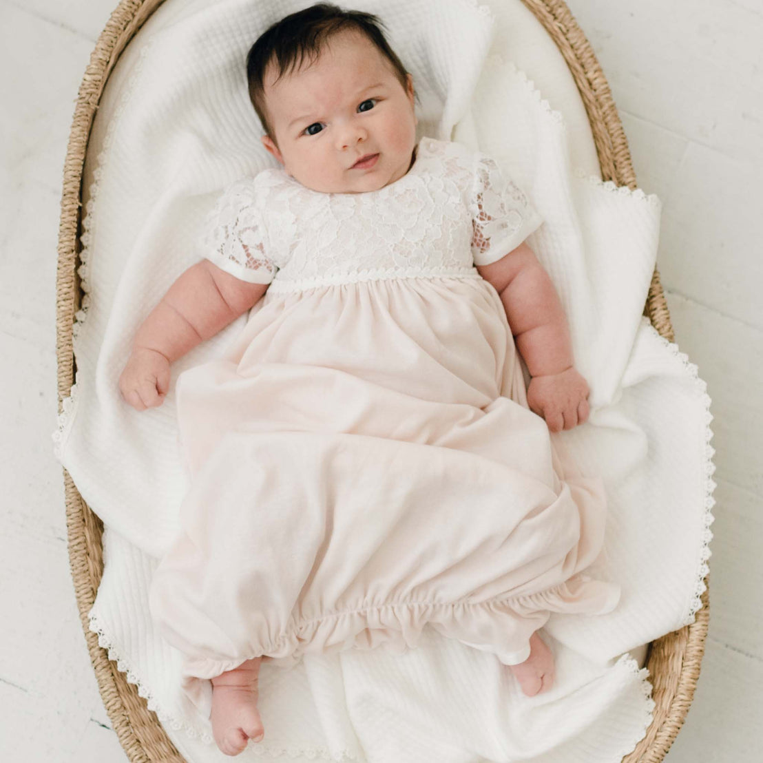 A content Rose Layette & Bonnet baby in a traditional pink dress with lace details lying in a woven basket, looking directly at the camera with a subtle smile.